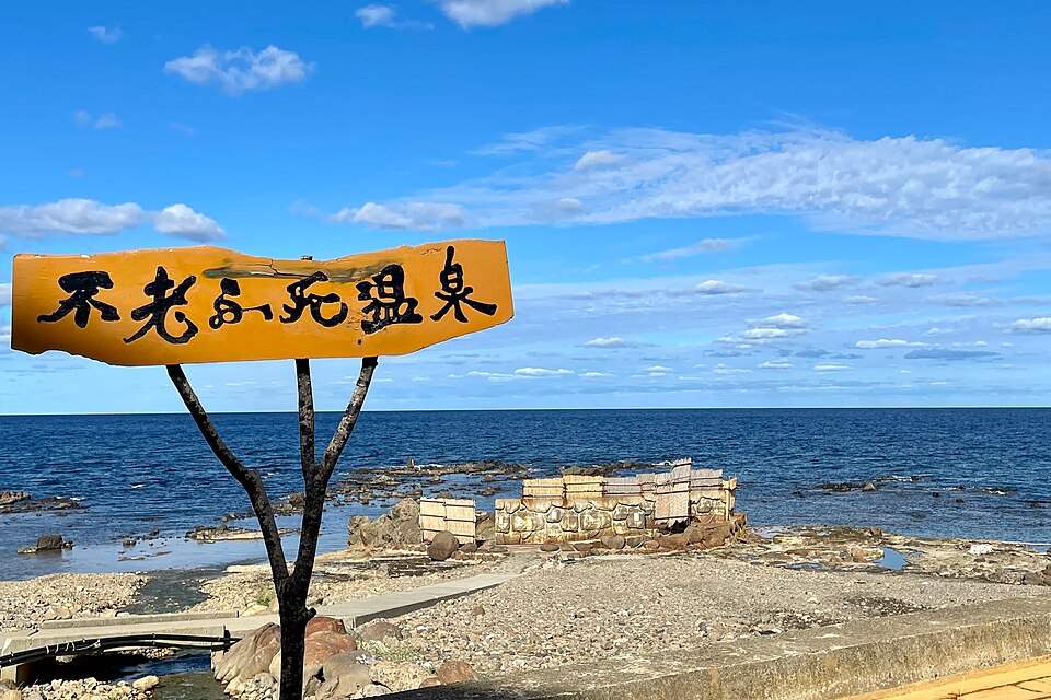 黄金崎不老ふ死温泉の看板と、日本海の波打ち際にある露天風呂